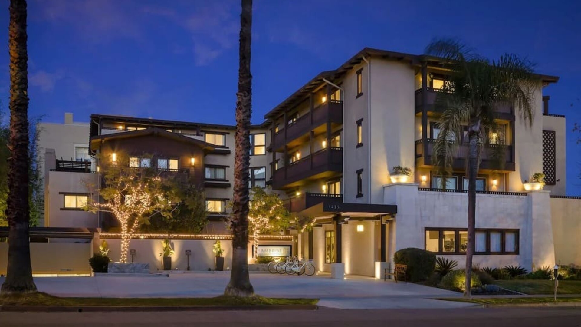 Exterior view at dusk with lit facade, palm trees, and entrance canopy along the street at The Ambrose Hotel Santa Monica.