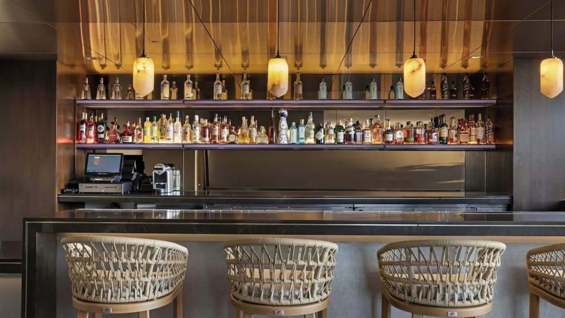 Sleek bar with backlit shelves, bottle display, pendant lighting, and woven bar stools at AC Hotel By Marriott Beverly Hills.