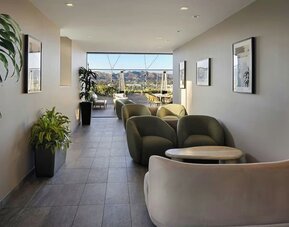 Lounge corridor with cushioned seating, small tables, plants, and open view toward outdoor terrace at AC Hotel By Marriott Beverly Hills.