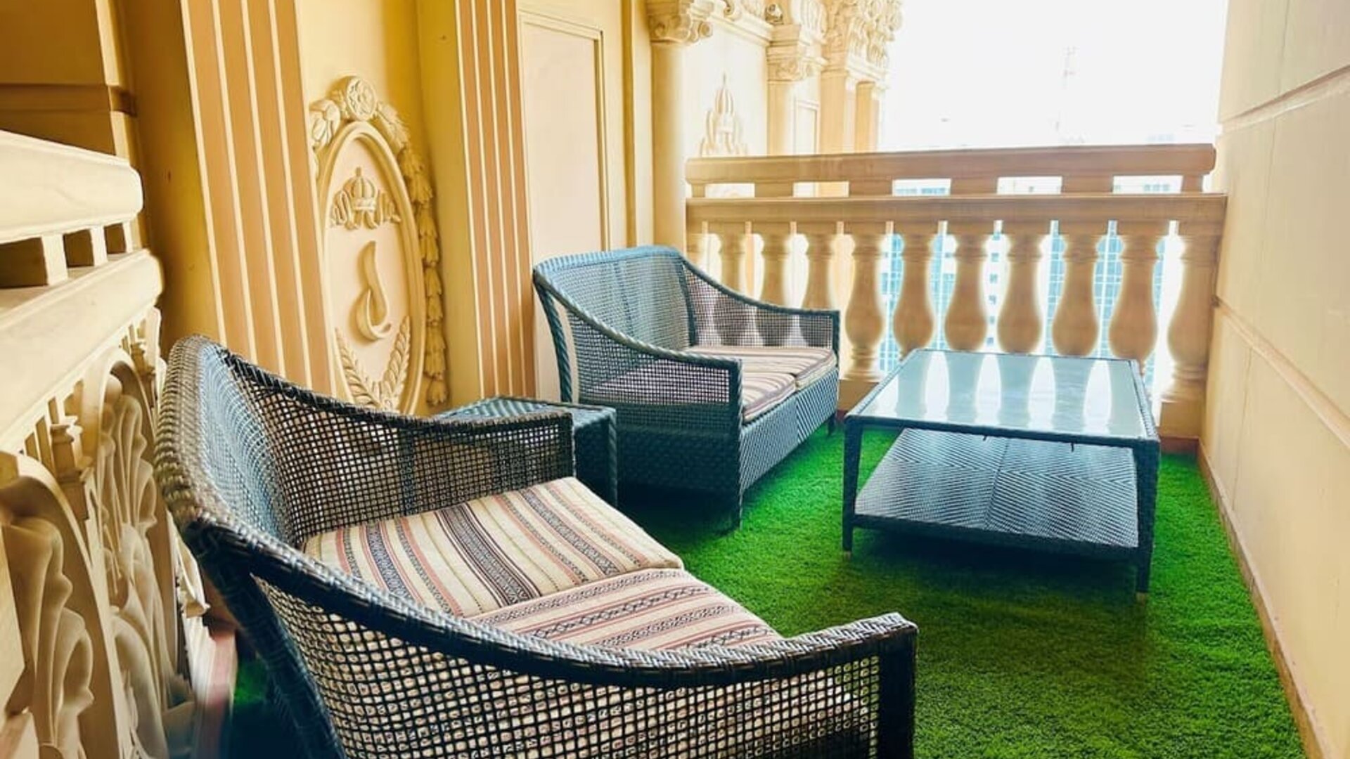 Narrow balcony with two wicker armchairs and a glass table on green turf, framed by ornate cream railings