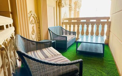 Narrow balcony with two wicker armchairs and a glass table on green turf, framed by ornate cream railings