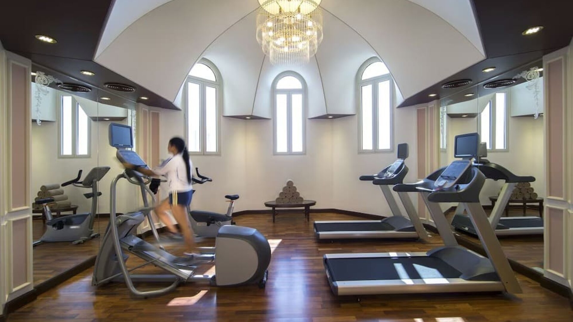 Bright fitness room with treadmills, elliptical machines, mirrored walls, and arched windows overhead