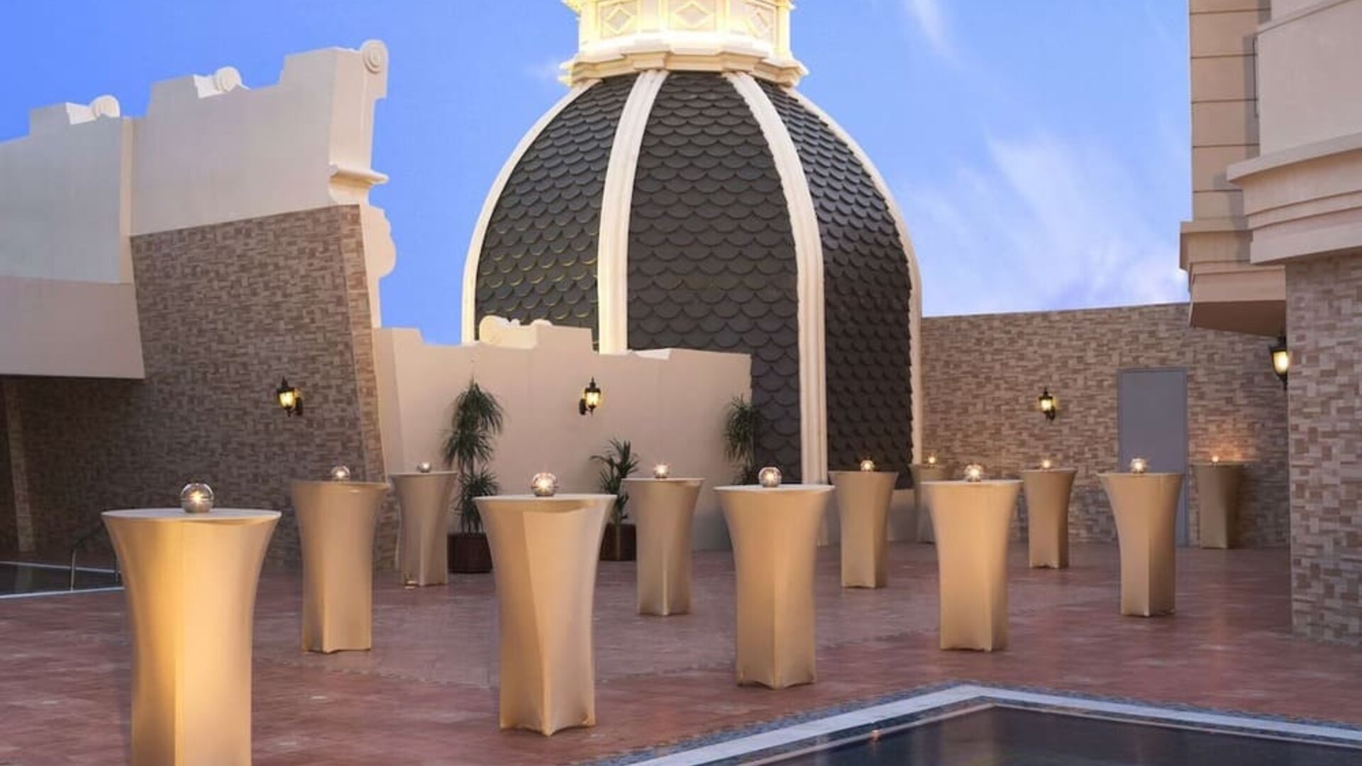 Rooftop terrace with small cocktail tables, domed structure, and reflecting pool under clear sky