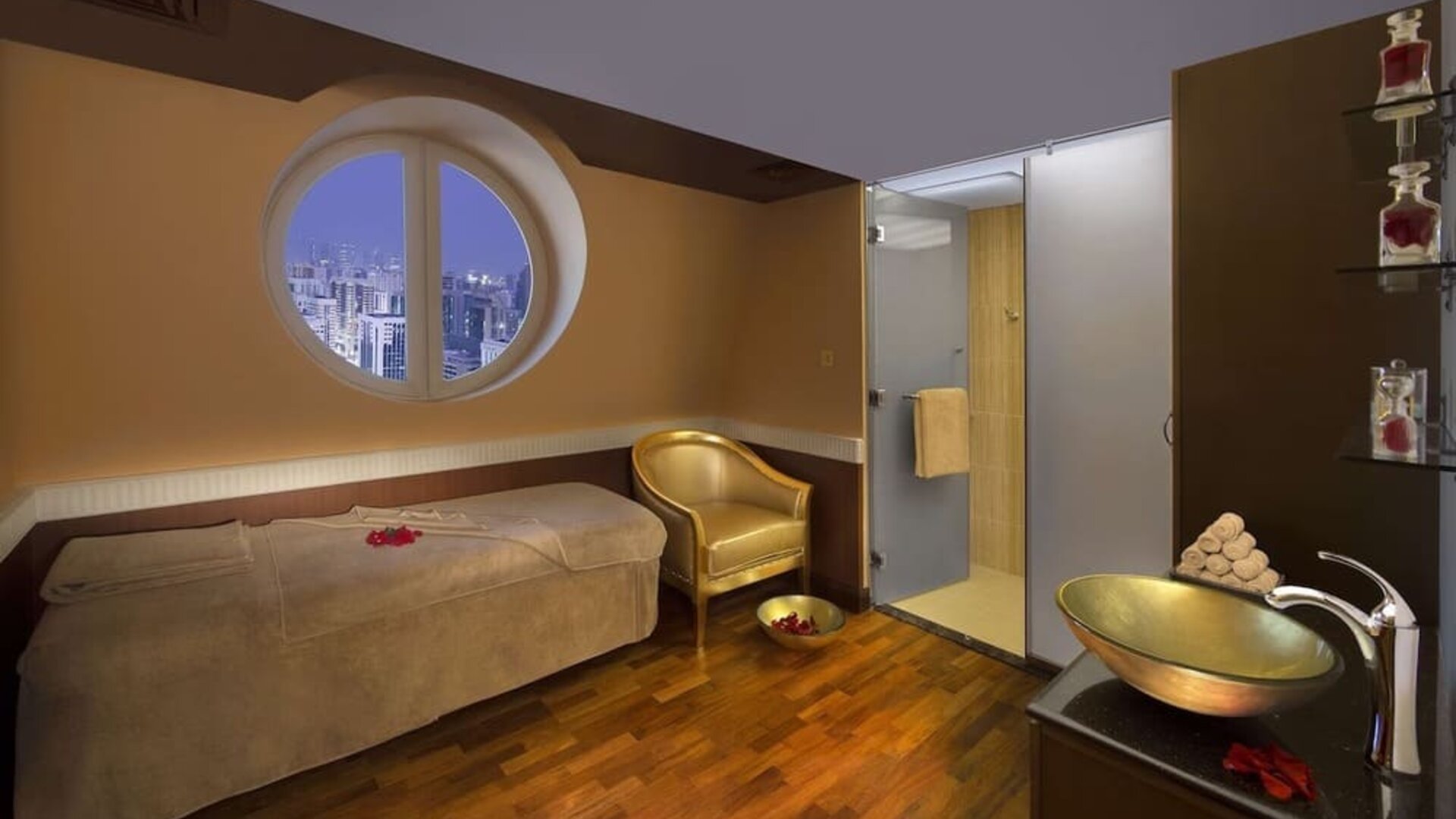 Spa treatment room with massage bed, round window, glass shower, and wooden flooring