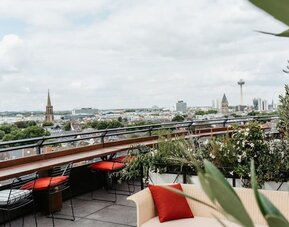 Rooftop terrace with cushioned seating, potted plants, and wide city skyline views beyond glass railing