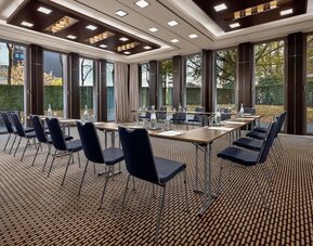 Meeting room with U-shaped tables, blue chairs, large windows, and garden view outside