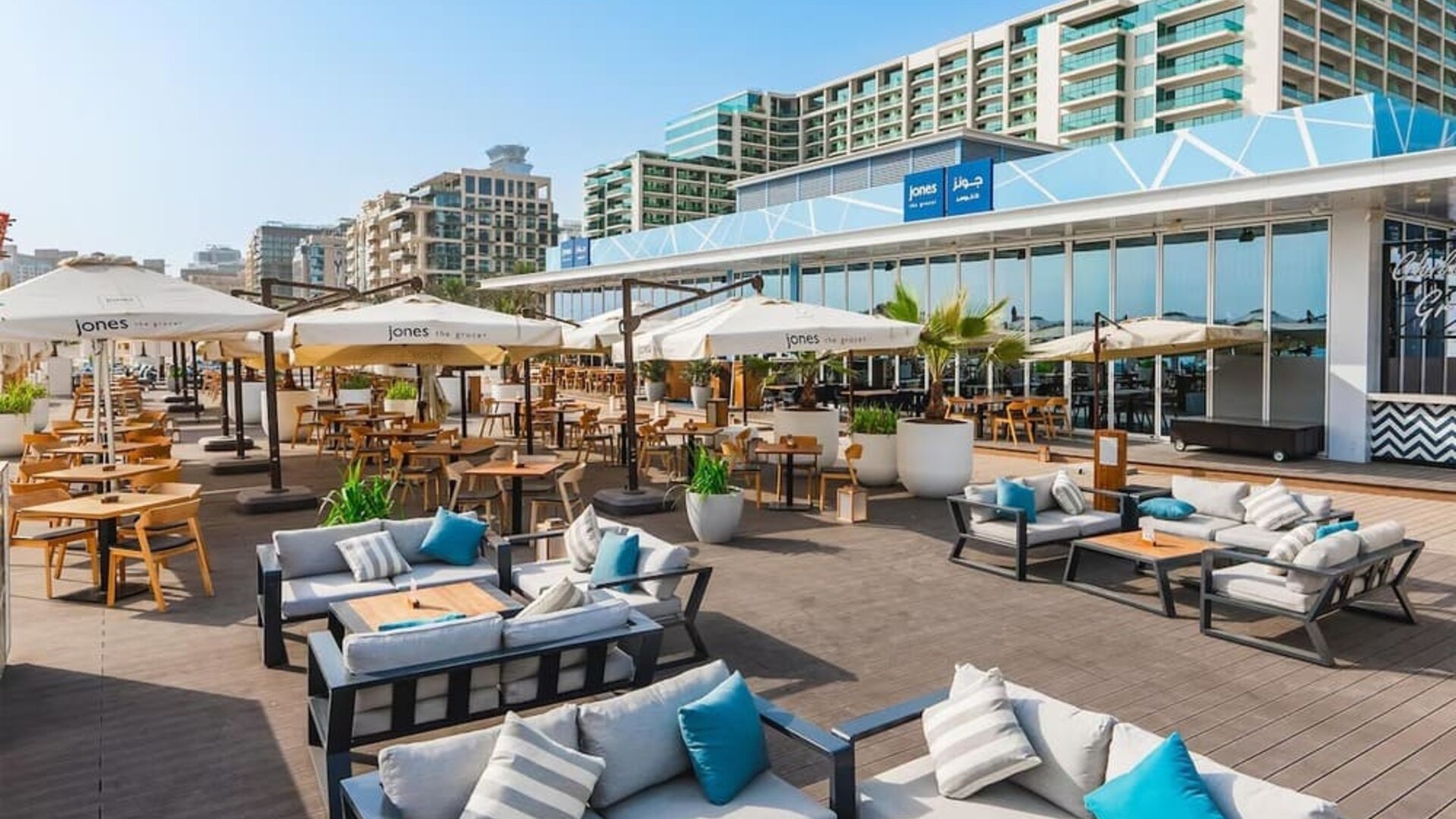 Open-air terrace with cushioned seating, dining tables, umbrellas, and low-rise buildings in background