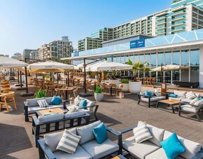 Open-air terrace with cushioned seating, dining tables, umbrellas, and low-rise buildings in background