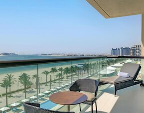 Balcony with lounge chair and table overlooking palm-lined beach, turquoise water, and distant city skyline