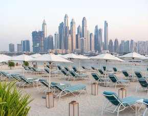 Sandy beach with rows of teal lounge chairs and white umbrellas facing calm water and tall city skyline