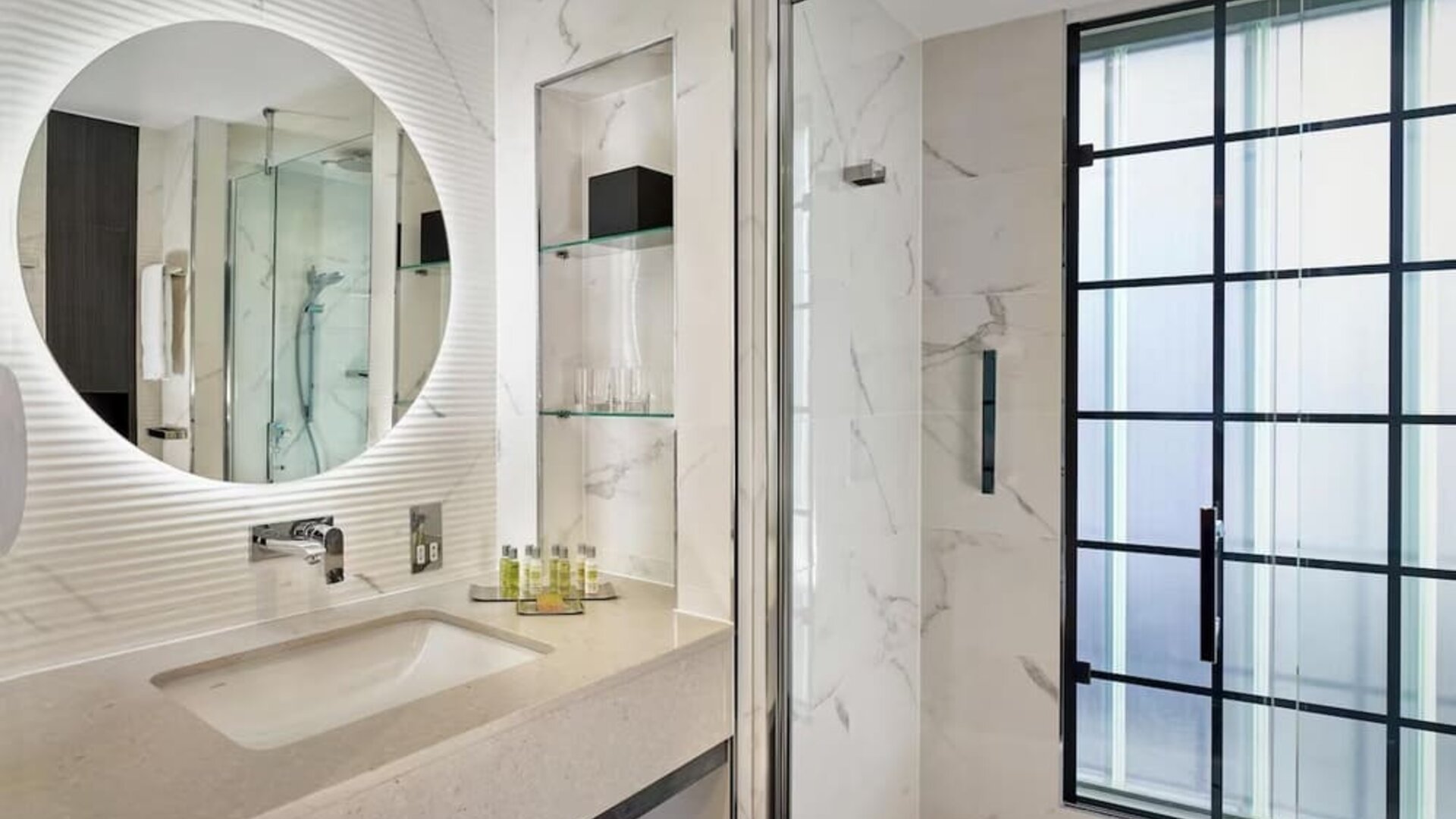 Marble bathroom with glass shower, round mirror above sink, and black-framed frosted window letting in soft light