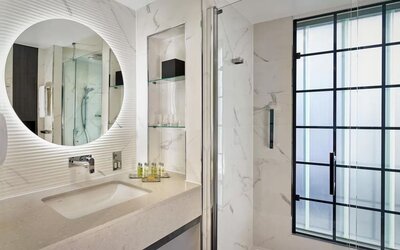 Marble bathroom with glass shower, round mirror above sink, and black-framed frosted window letting in soft light