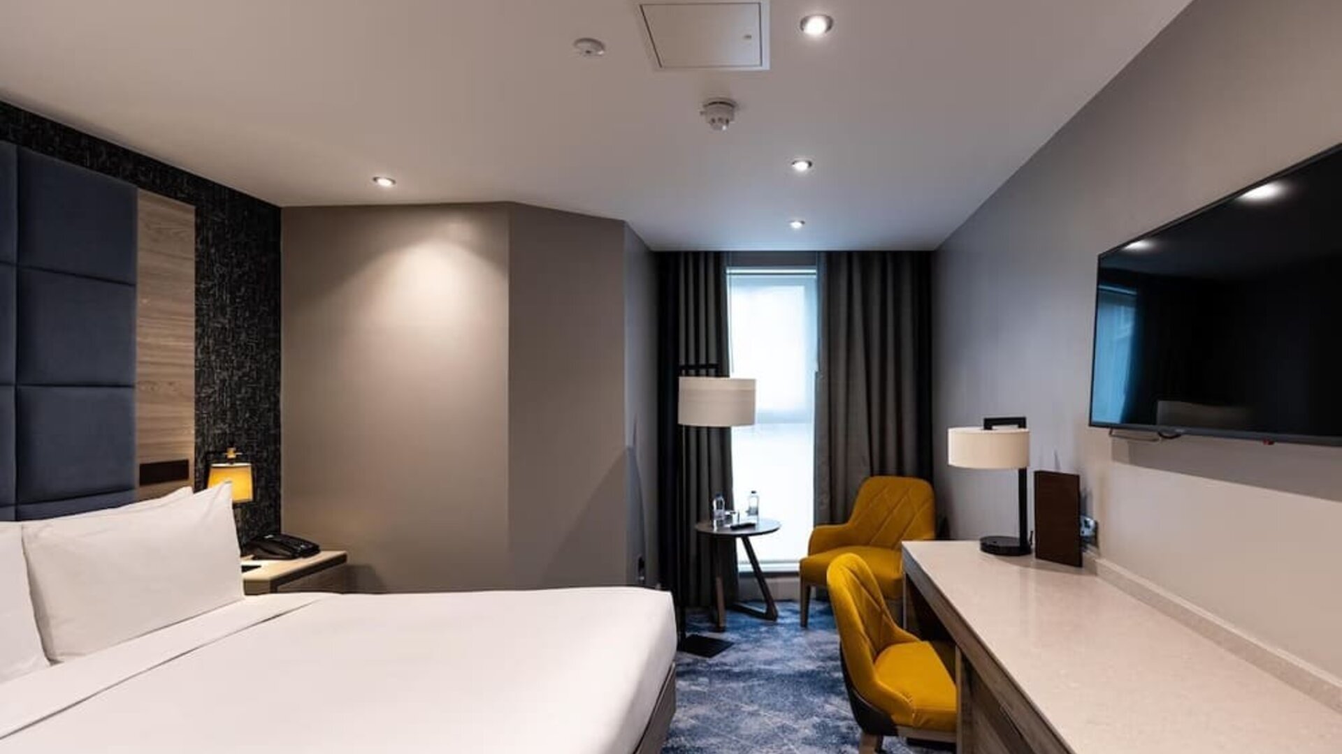 King bed beside long desk and wall-mounted TV, with yellow chair and floor lamp near a narrow window