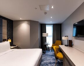 King bed beside long desk and wall-mounted TV, with yellow chair and floor lamp near a narrow window