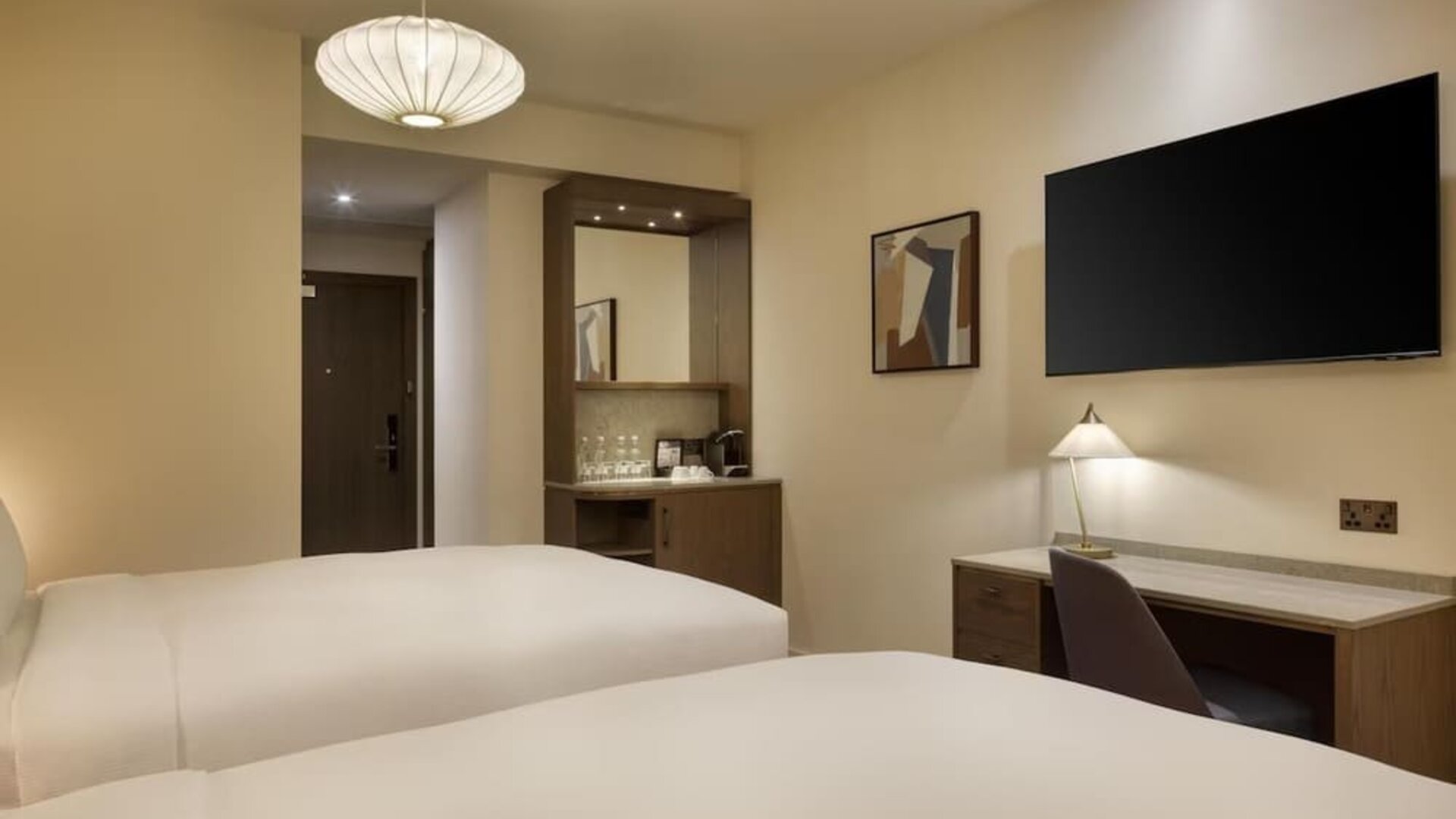 Two-bed guest room with desk, wall-mounted TV, and coffee station near entry in warm neutral tones