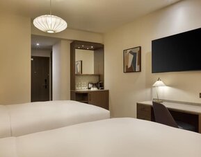Two-bed guest room with desk, wall-mounted TV, and coffee station near entry in warm neutral tones