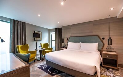 Spacious king room with large bed, padded headboard, desk, and seating area by tall window with dark curtains