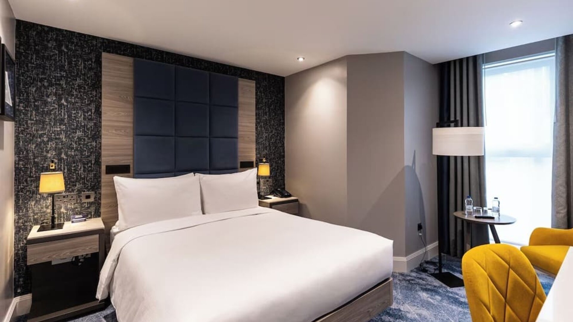 King bed with padded blue headboard, yellow chairs by window, and textured accent wall behind bedside tables