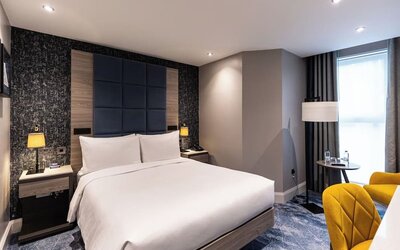 King bed with padded blue headboard, yellow chairs by window, and textured accent wall behind bedside tables