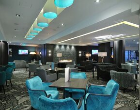 Modern lounge with teal chairs, patterned carpet, fireplace wall, and soft overhead lighting accents