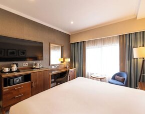 King guest room with wall-mounted TV, wood desk, blue accent chair, and large curtained window