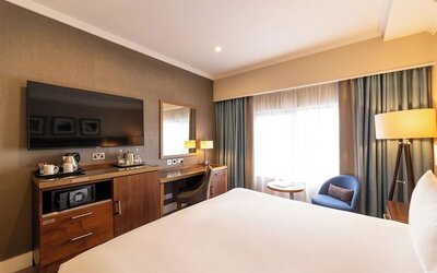 King guest room with wall-mounted TV, wood desk, blue accent chair, and large curtained window