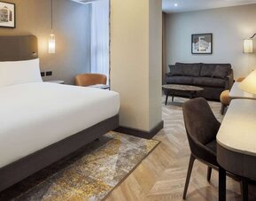 Suite-style king room with separate sitting area, sofa, curved desk, and modern lighting fixtures