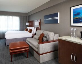 Spacious guest room with king bed, sofa seating area, ottoman, and kitchenette counter at Courtyard By Marriott Los Angeles LAX/Century Boulevard.