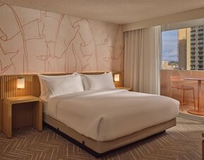 Guest room with king-size bed, modern wood accents, soft ambient lighting, and private balcony seating overlooking Waikiki at The Ambassador Hotel Of Waikiki