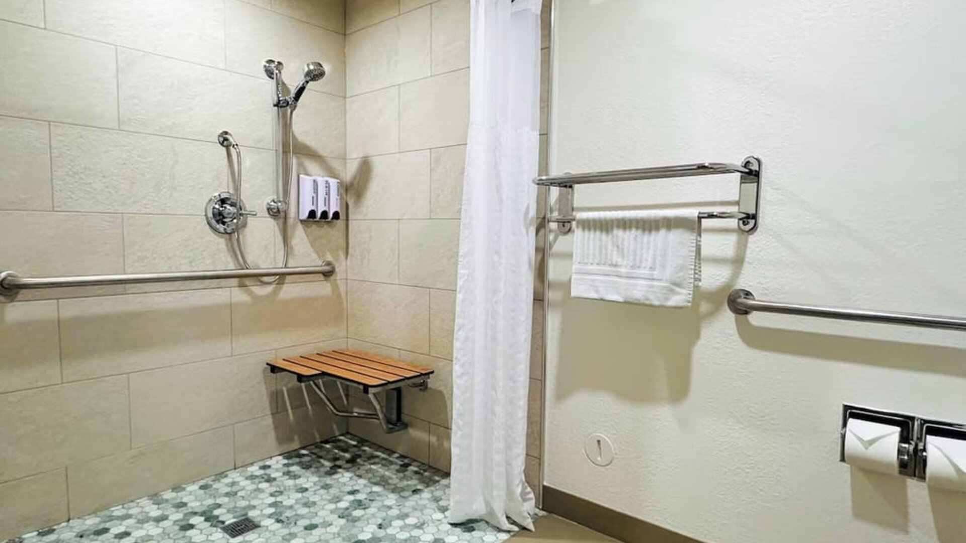 Accessible bathroom with roll-in shower, grab bars, fold-down bench, and tiled flooring at The Hills Inn Eagle Rock.