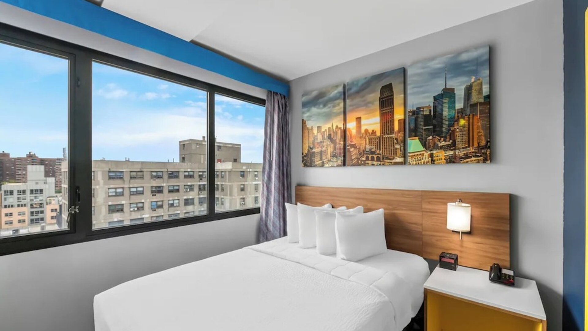 Queen bed beside large city-view window with skyline artwork and bright accents at Days Inn Chinatown NYC.