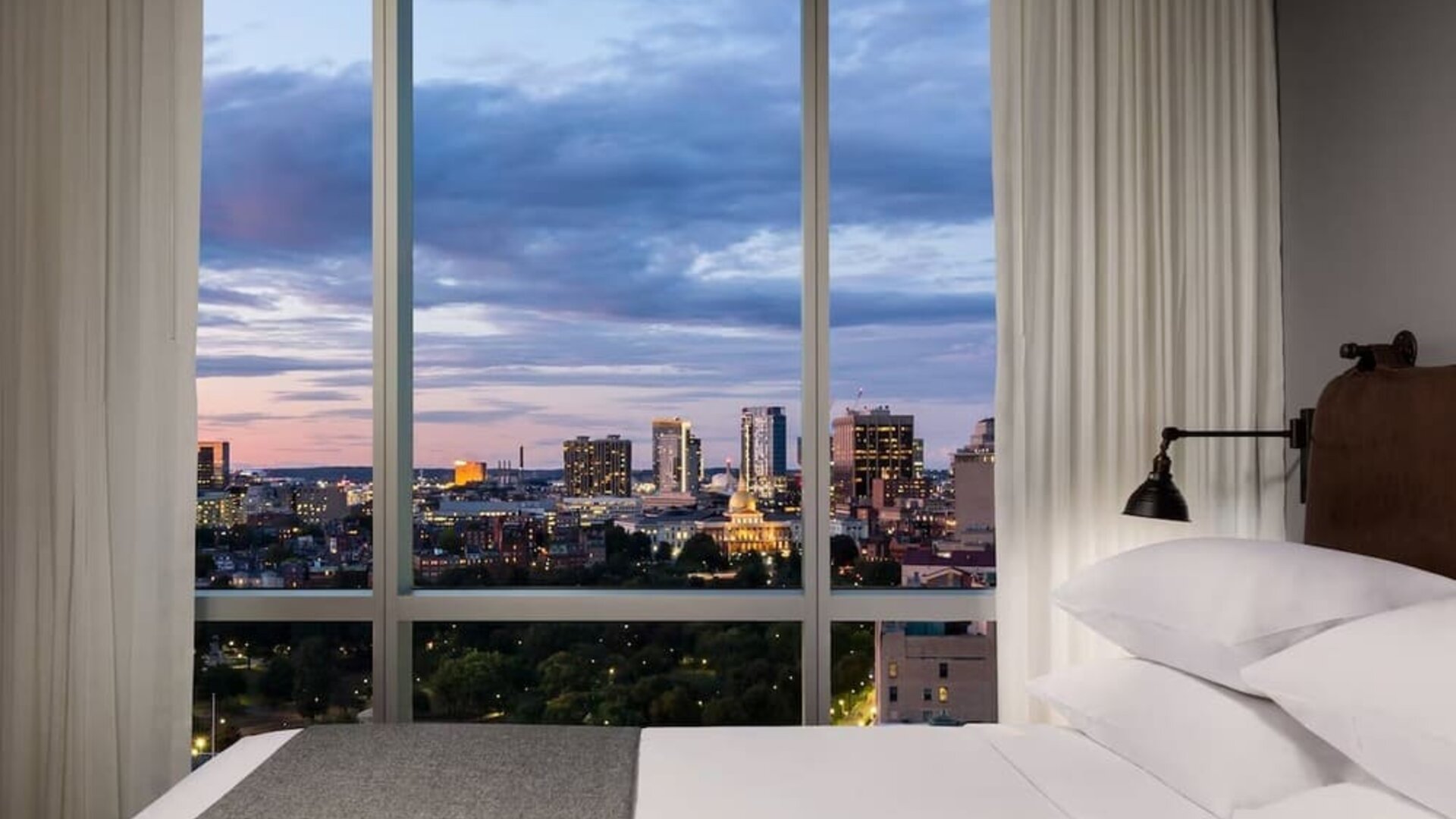Bedroom with floor-to-ceiling windows showcasing evening skyline views at Moxy Boston Downtown.