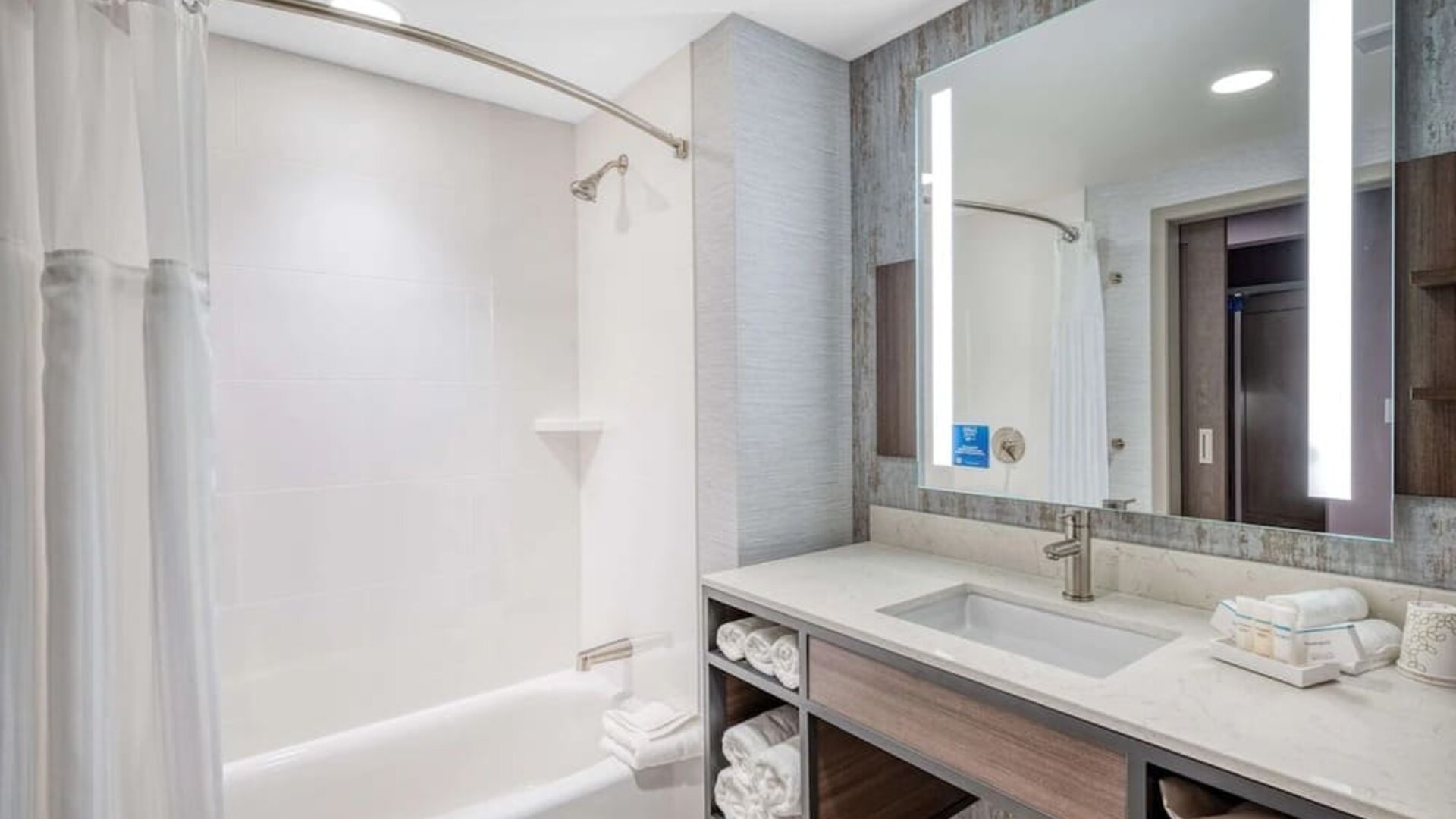 Modern bathroom with illuminated mirror, bathtub shower combo, and marble-style vanity at Hilton Garden Inn Boston Logan Airport.