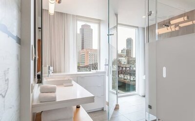 Sleek modern bathroom with floor-to-ceiling windows, glass shower enclosure, and stunning Boston waterfront views at The Envoy Hotel, Autograph Collection.