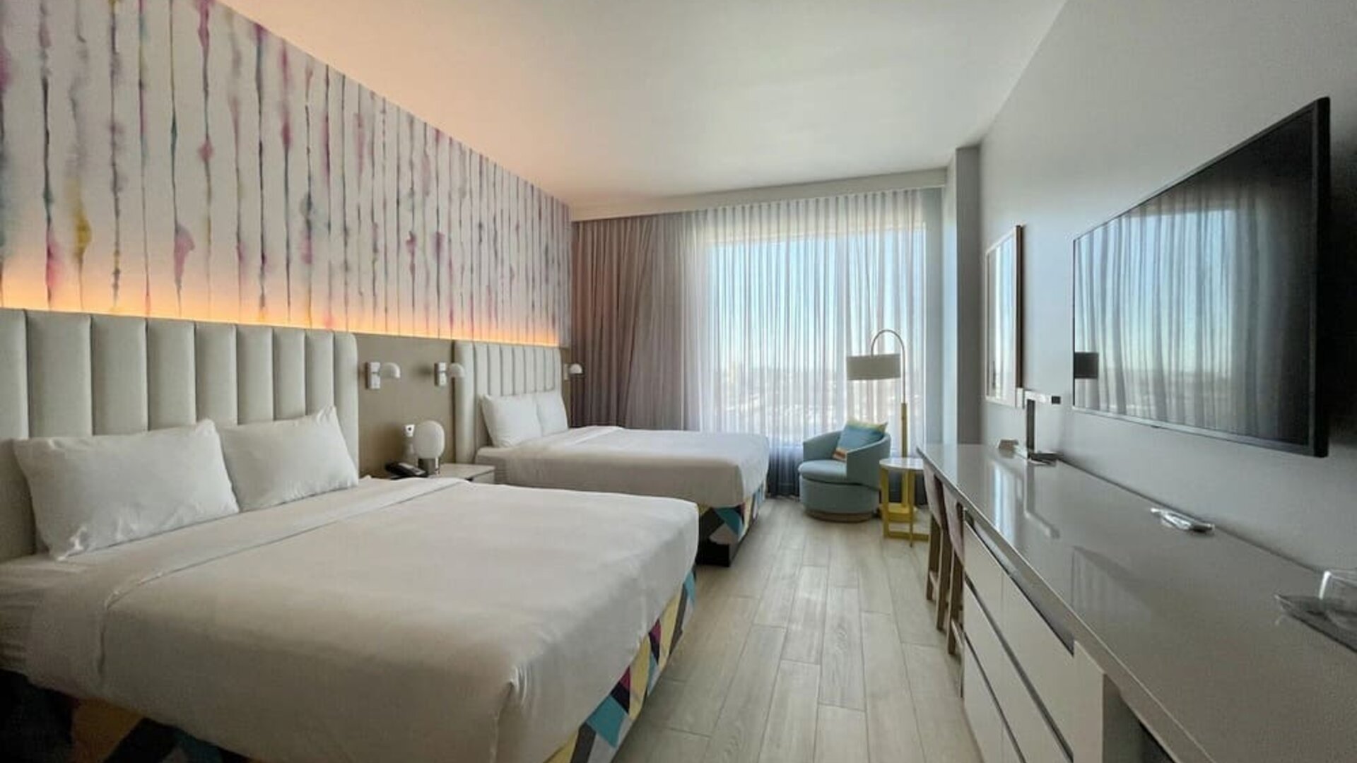 Spacious guest room with two beds, modern furnishings, oversized television, work area, and large windows providing abundant natural light.