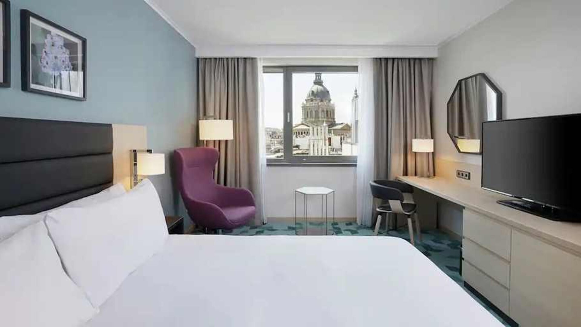 lovely kind bedroom with TV, workspace, couch, and city views at Hilton Garden Inn Budapest City Centre.