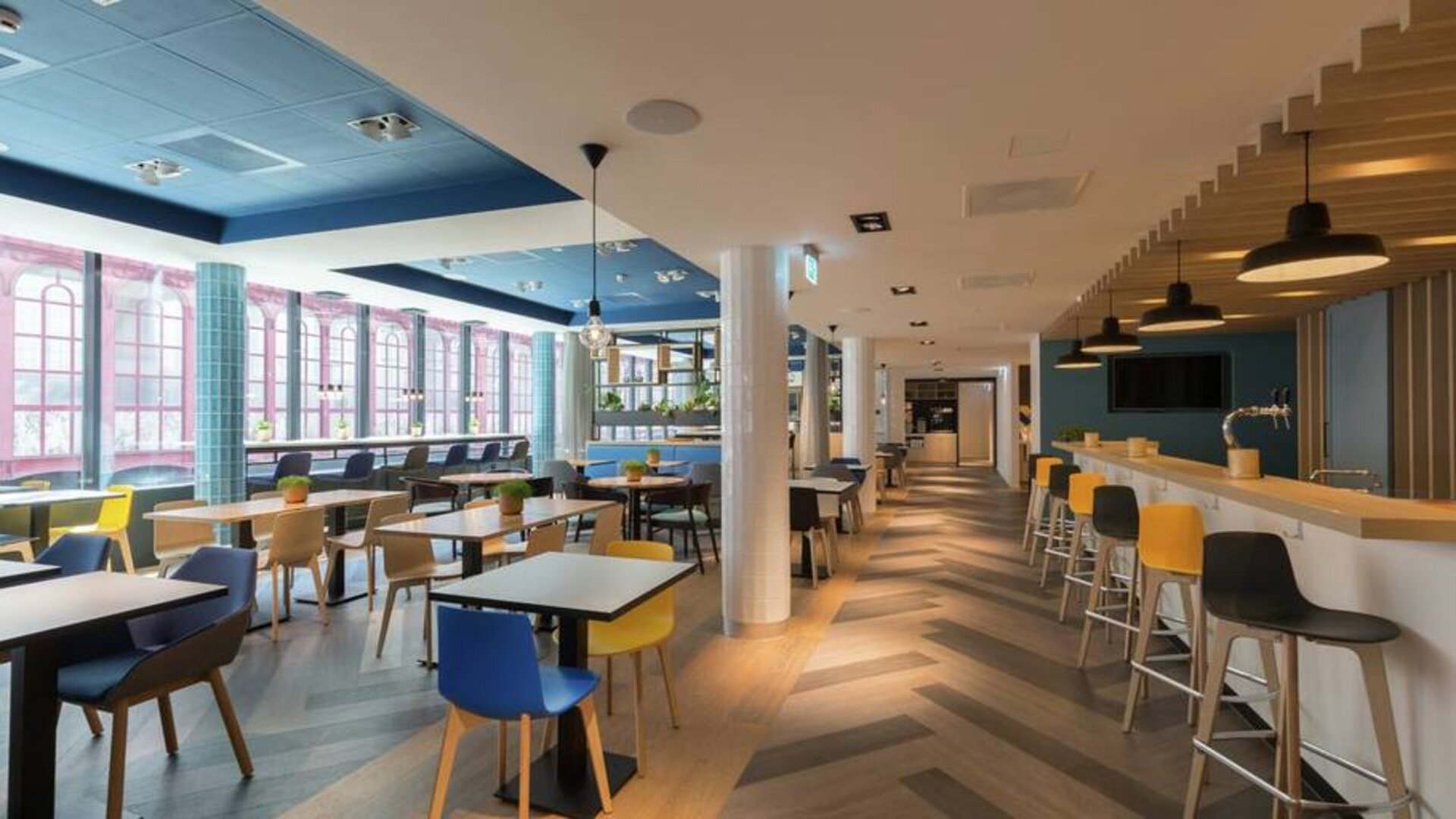Large lobby with tall ceiling and floor to ceiling window view of downtown Antwerp, various types of seating for socializing, working, eating.