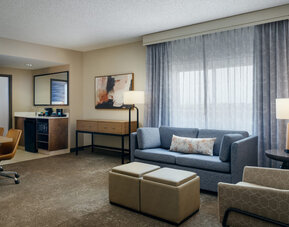 Spacious suite living area with comfortable sofa seating, work space, mini refreshment station, and large windows providing natural light at Embassy Suites By Hilton Charleston Airport Convention Center.