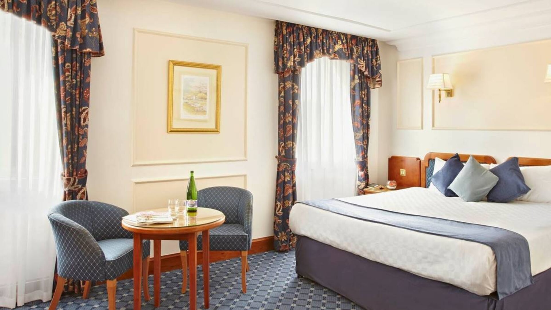 An executive double standard room at the Thistle London Heathrow Terminal 5, with large bed, windows, and coffee table and chairs.