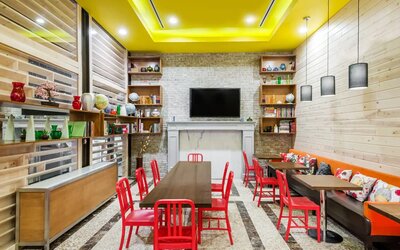 A long rectangular wooden table in the middle with red chairs. Several other smaller tables for 4 to work, eat, socialize at. Yellow lit ceiling and white fireplace mantle frames the room.