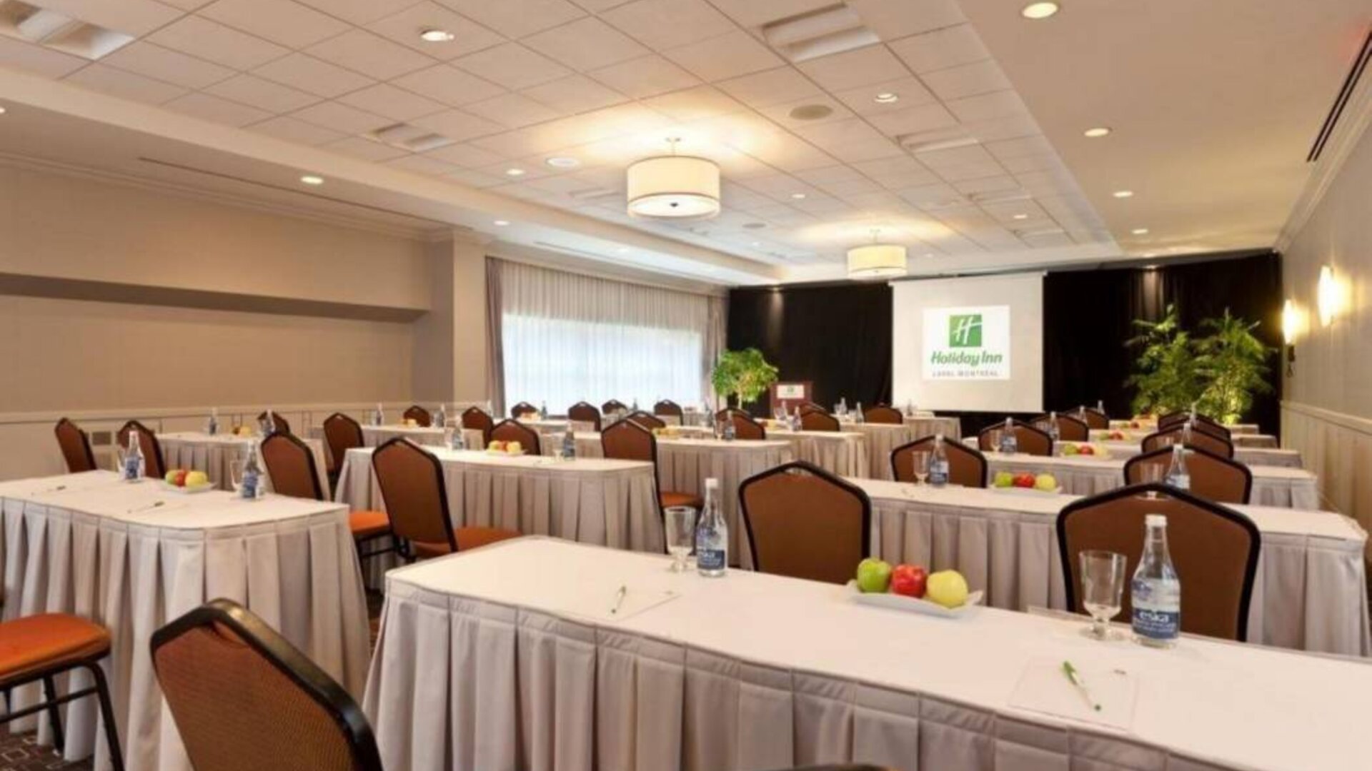 Meeting room with numerous tables and seating arranged to face a lectern, beside which is a projector screen.
