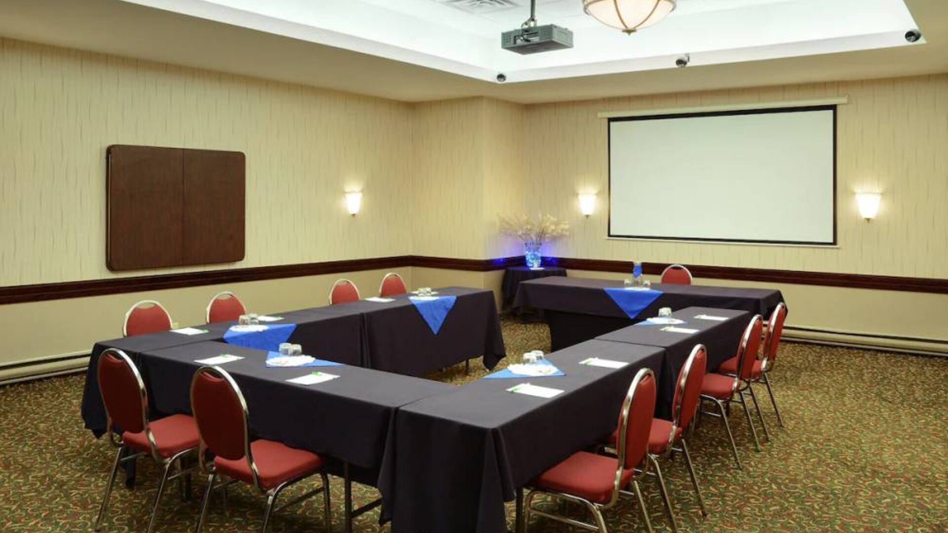 Meeting room with numerous tables, projector screen, and seating for around a dozen.