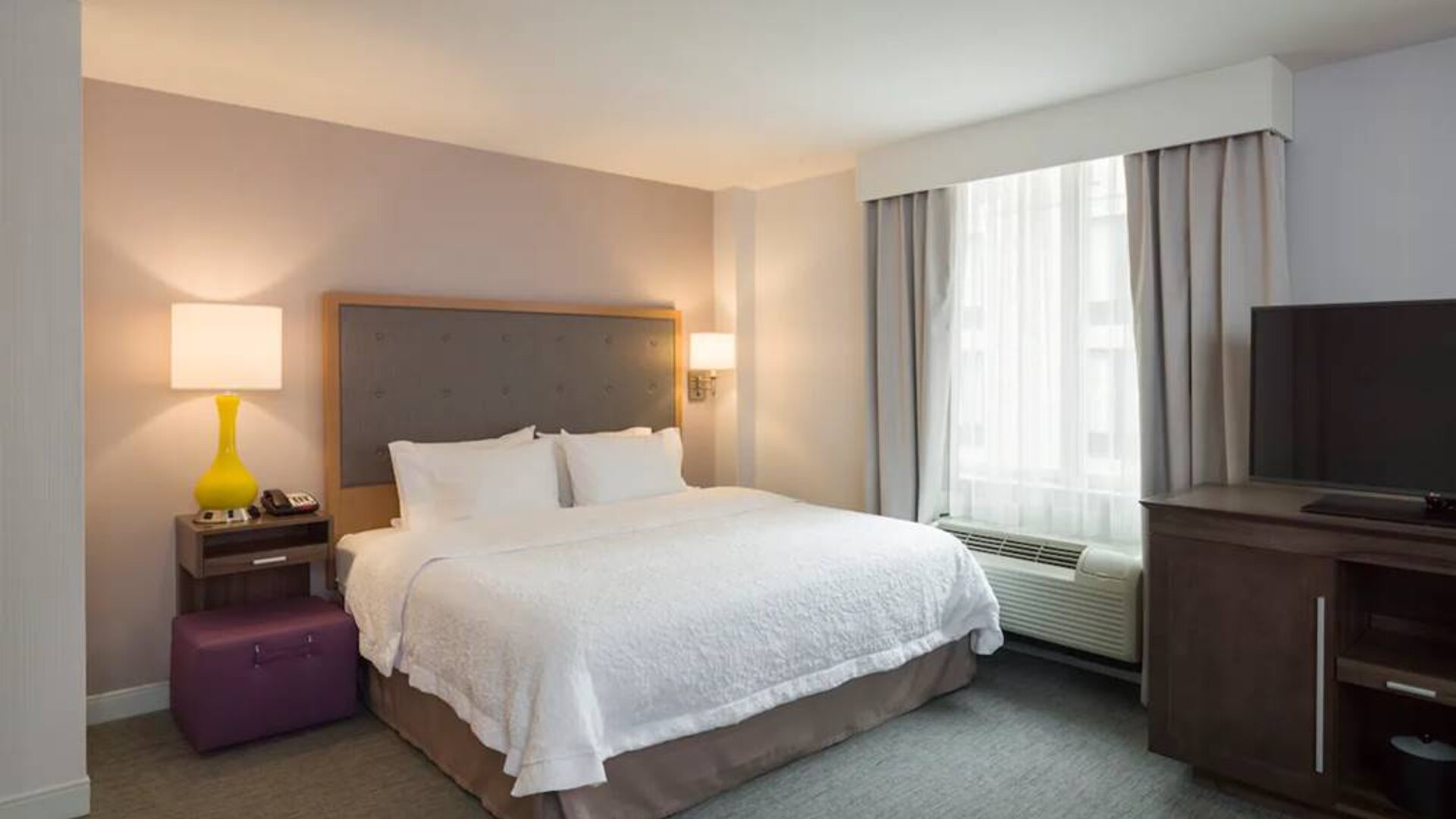 One of the Hampton Inn Times Square South’s king bed guest rooms, with window, TV, and bedside table.