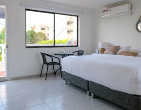 A bright king bedroom with white bedding, beige accent cushions, a round wall mirror, a woven bedside lamp and a small table with two chairs beside the window, with access to an outdoor balcony at Mucura Hotel And Spa.