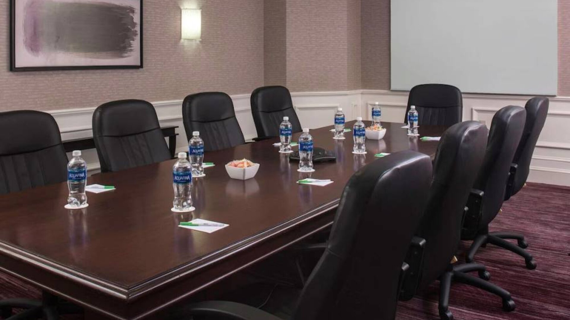 Sonesta Select Boston Lowell Chelmsford meeting room, including long wooden table, nine leather swivel chairs, and a projector screen.
