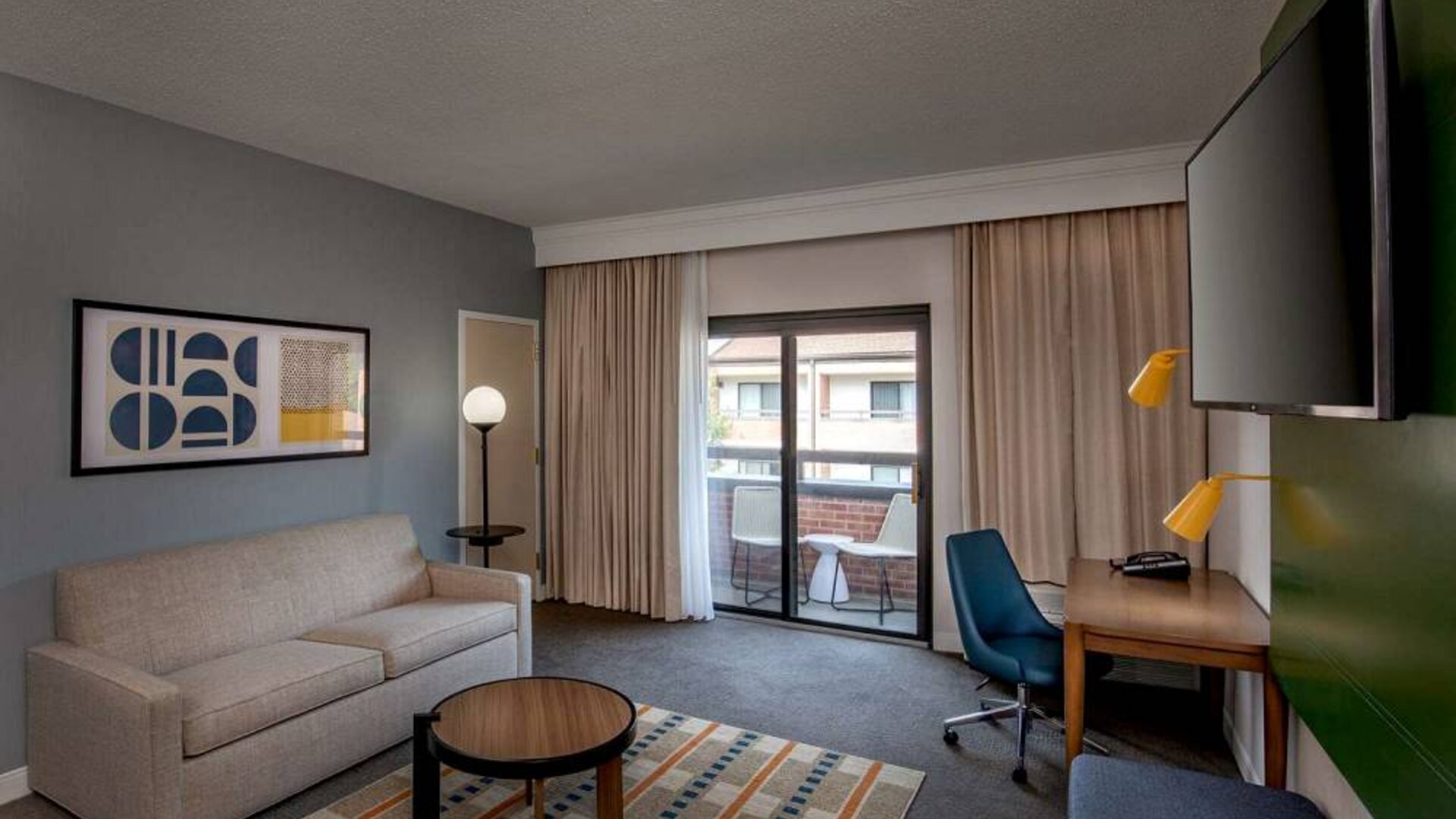 Sonesta Select Boston Foxborough Mansfield guest room lounge, furnished with sofa, desk, coffee table, and TV, with a balcony outside.