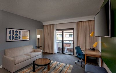 Sonesta Select Boston Foxborough Mansfield guest room lounge, furnished with sofa, desk, coffee table, and TV, with a balcony outside.