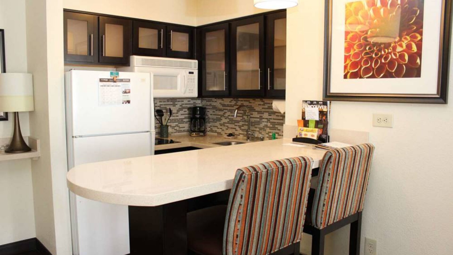 Guest room kitchen in Sonesta ES Suites Portland Vancouver 41st Street, featuring fridge-freezer, microwave, breakfast bar and two stools.