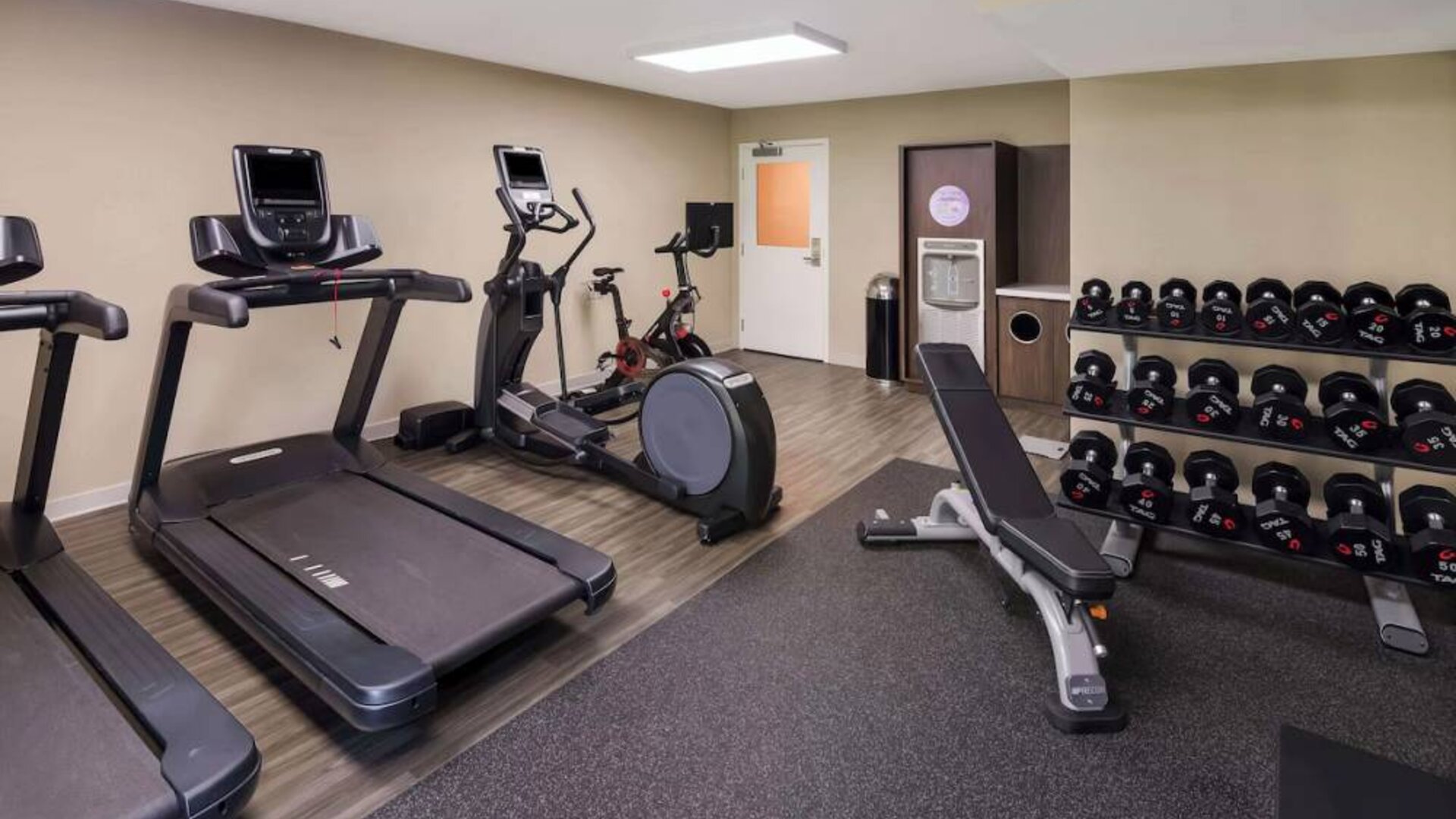 The fitness center in Sonesta ES Suites Andover Boston is equipped with rows of free weights, a bench, and a variety of exercise machines.