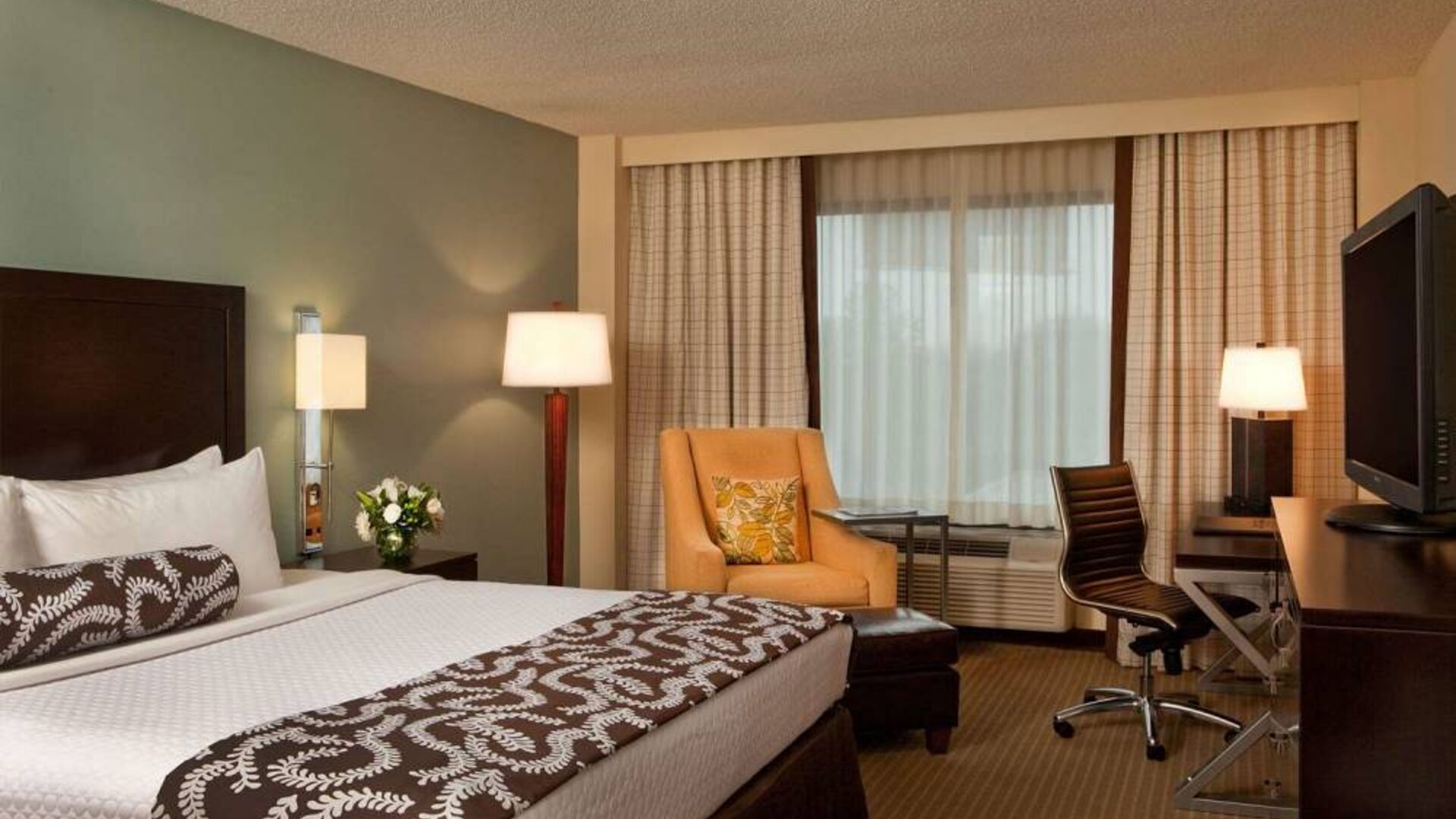 Double bed guest room in Sonesta Atlanta Airport North, featuring TV, window, armchair, and workspace desk and chair.